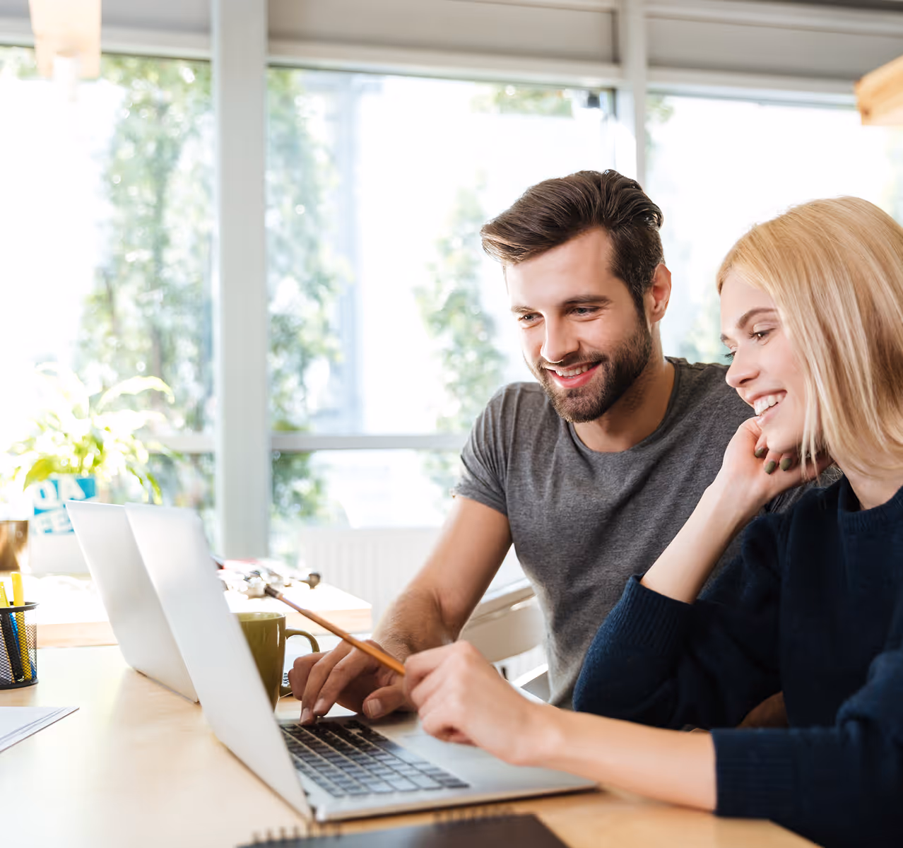 Deux collègues souriants travaillent ensemble sur un ordinateur portable dans un bureau lumineux.