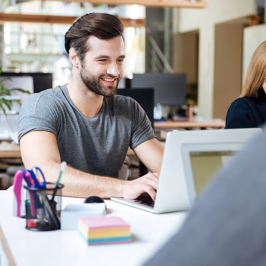 Un homme souriant utilise un ordinateur portable dans un bureau moderne avec des fournitures de bureau colorées sur la table.