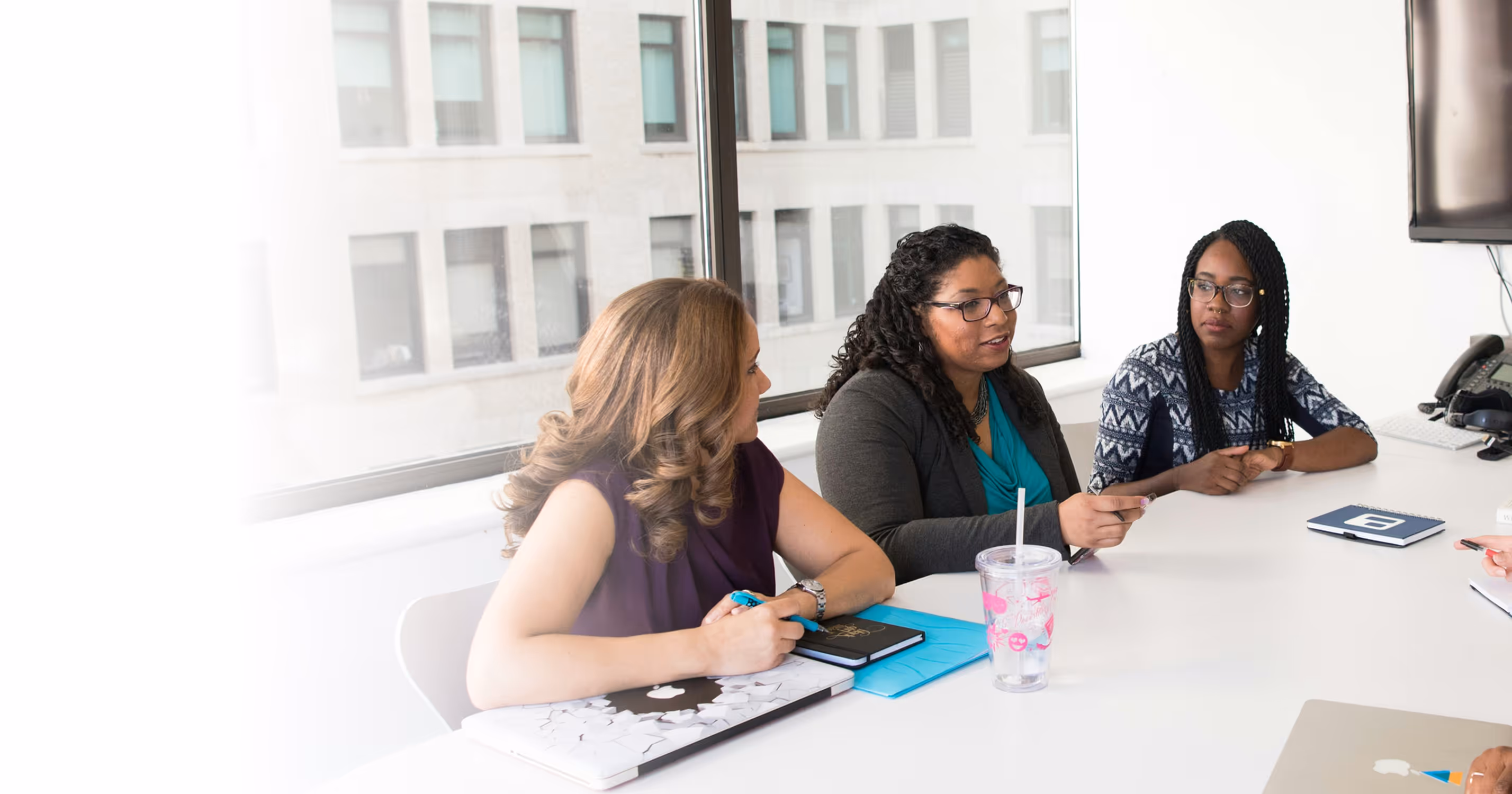 Trois femmes discutent autour d'une table dans une salle de réunion avec de grands fenêtres, avec des ordinateurs portables et carnets devant elles.