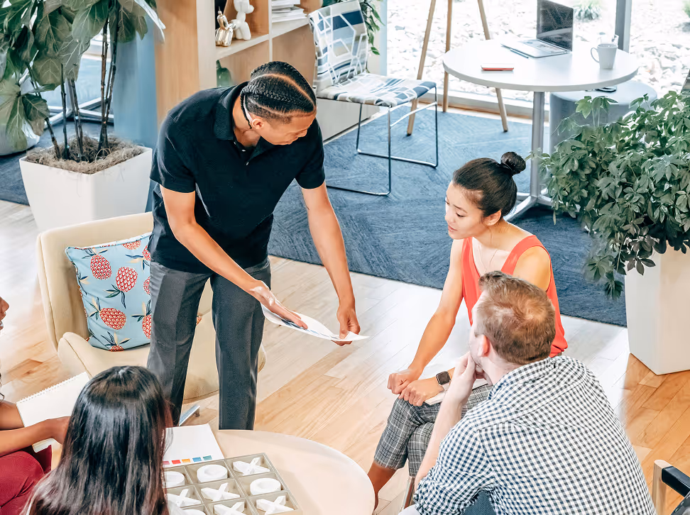 Un homme debout montre des documents à un groupe de personnes assises dans un bureau lumineux avec des plantes.