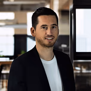 Portrait d'un homme souriant portant une veste noire et un t-shirt blanc dans un bureau moderne.