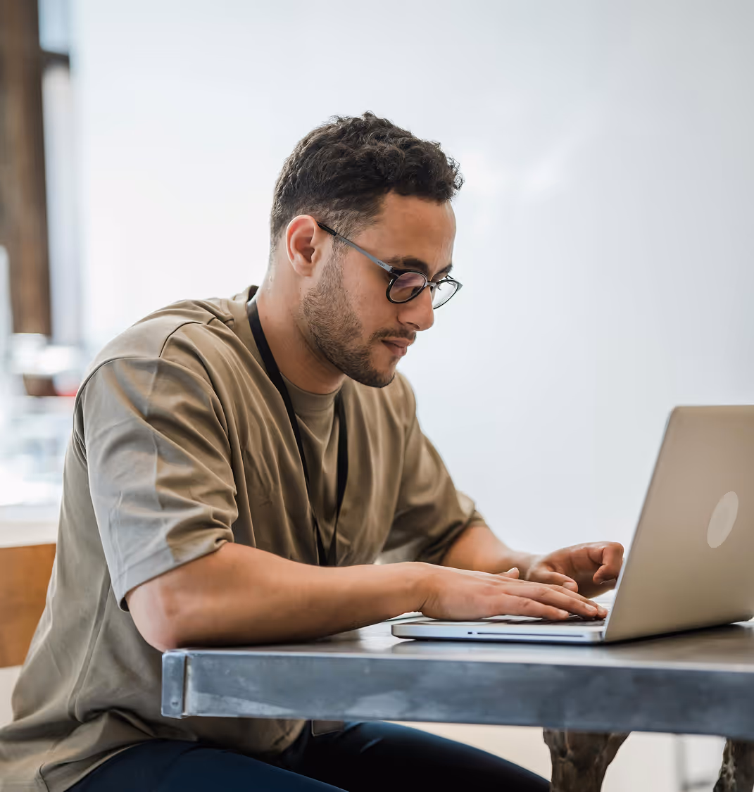 Jeune homme portant des lunettes et un t-shirt beige, travaillant sur un ordinateur portable à une table.