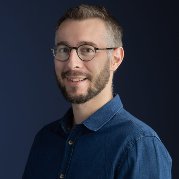 Homme portant des lunettes, chemise bleue, souriant devant un fond sombre.