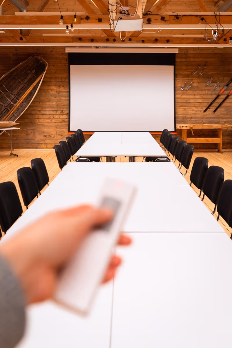 Hand holding a remote pointed at a blank projection screen in a conference room with long tables and black chairs.