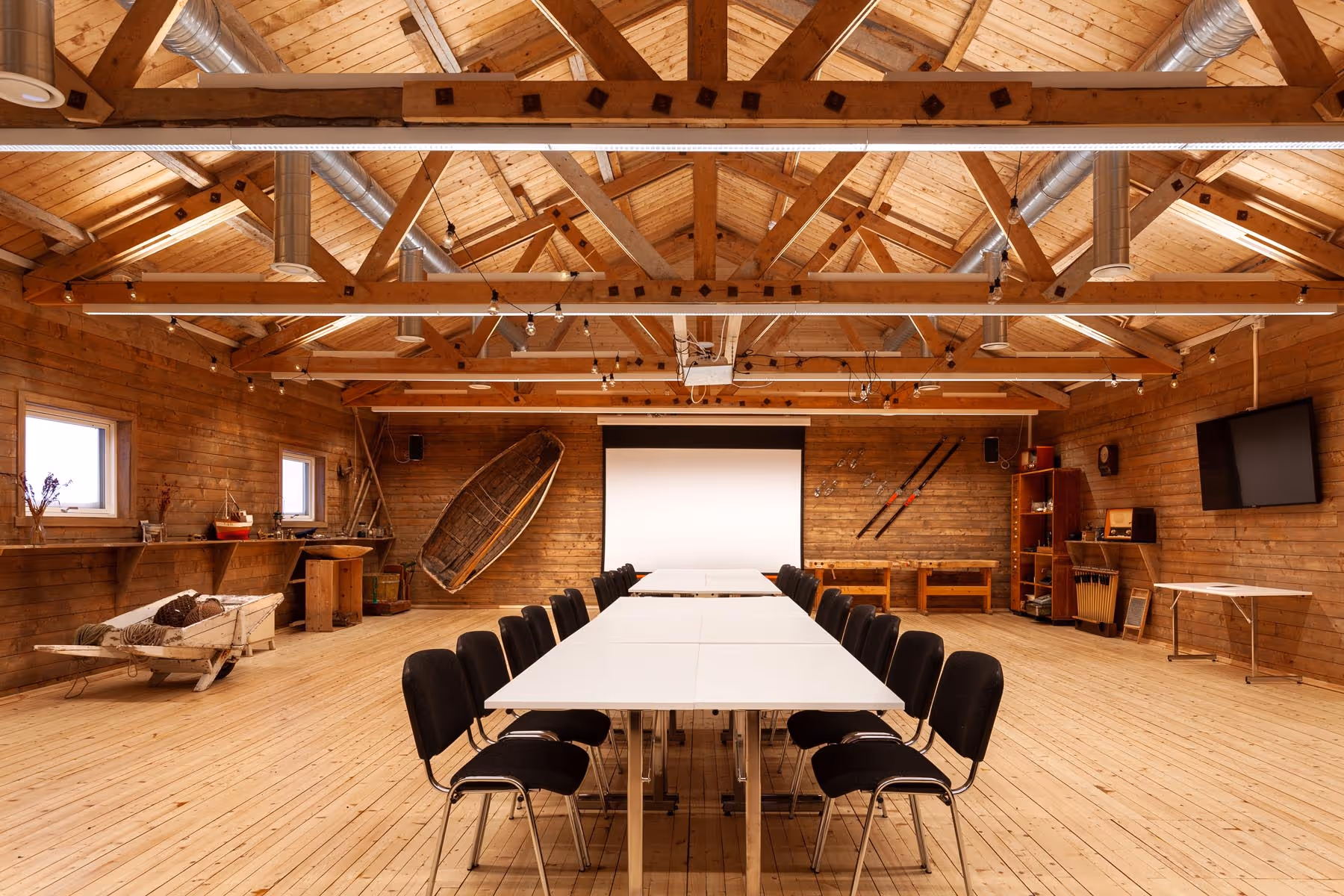 Rustic conference room with wooden walls and ceiling beams, white tables arranged in a U-shape, black chairs, and nautical decor including a small boat and oars.