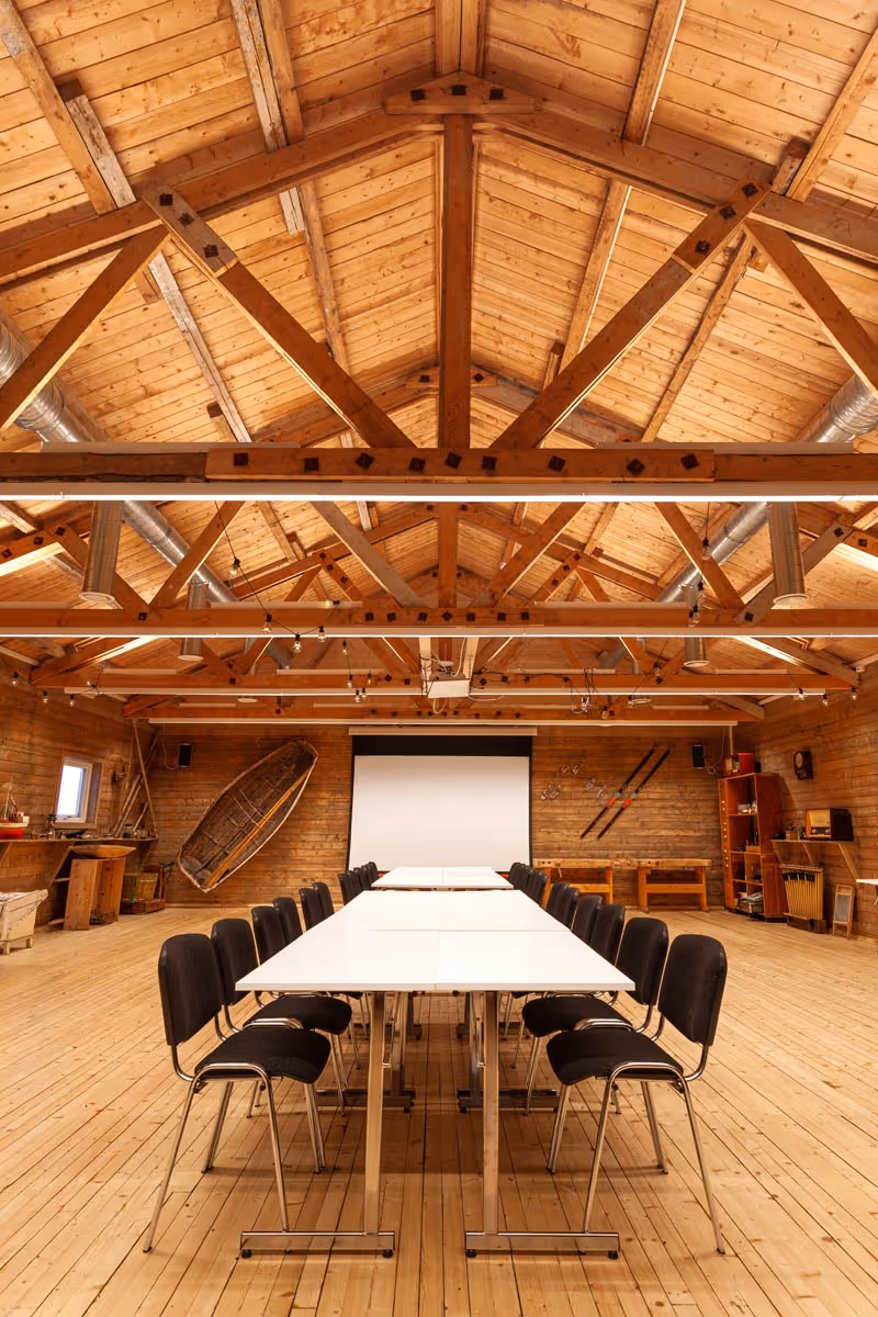 Long white tables arranged in a U shape with black chairs inside a wooden room with exposed beam ceiling and a projector screen.