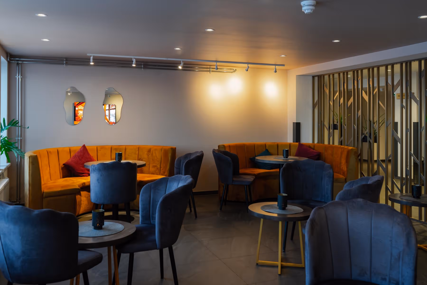 Modern lounge area with mustard yellow curved sofas, navy blue velvet chairs, round tables, and decorative mirrors on a gray wall.