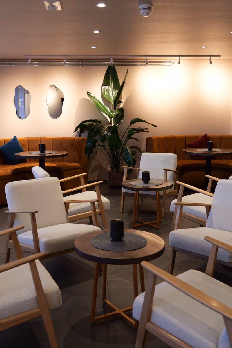 Modern lounge area with beige cushioned wooden armchairs around round wooden tables, brown curved sofas, and a large green plant against a cream wall with decorative mirrors.