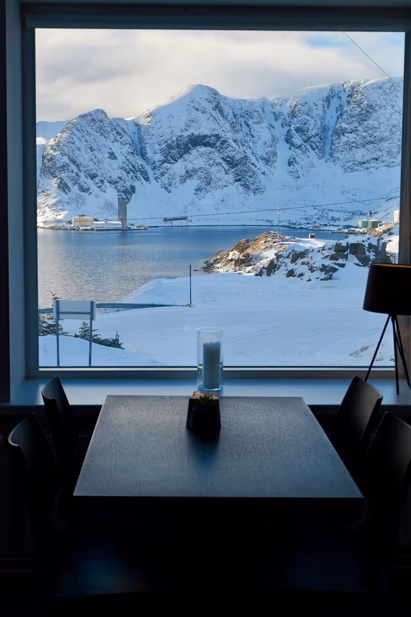 Snow-covered mountains and a calm lake viewed through a large window above a dark dining table with a candle and small plant.