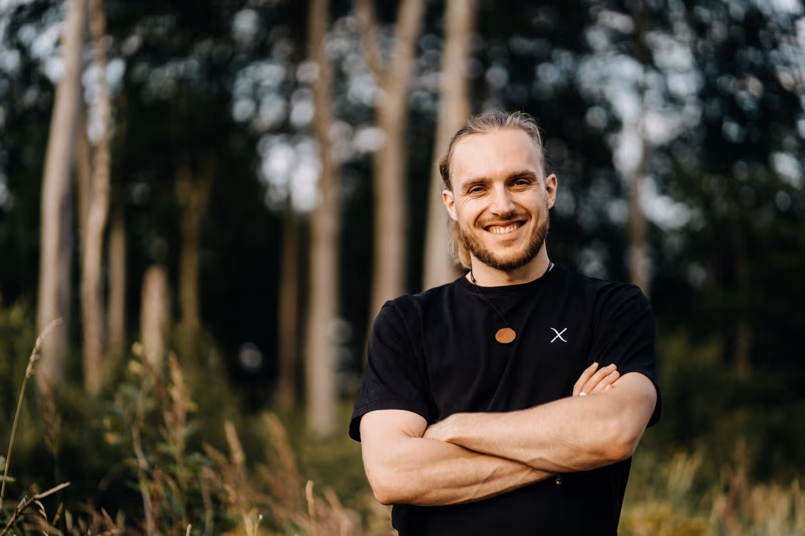 Foto des Gründers von Soulful Media Querformat - Junger Mann mit verschränkten Armen lächelnd zur Kamera in der Natur stehend vor Wald bei Sonnenuntergang