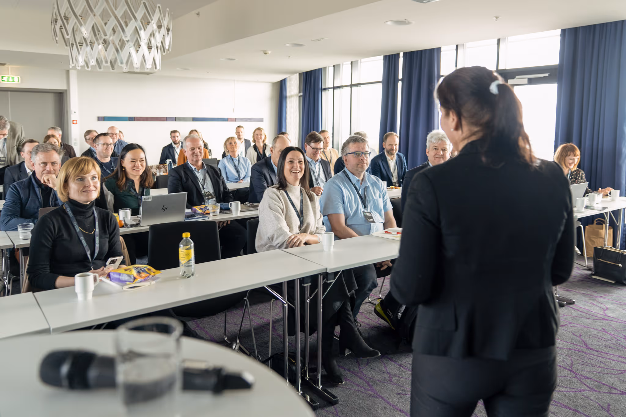 Audience of adults seated at tables attentively listening to a speaker in a conference room.