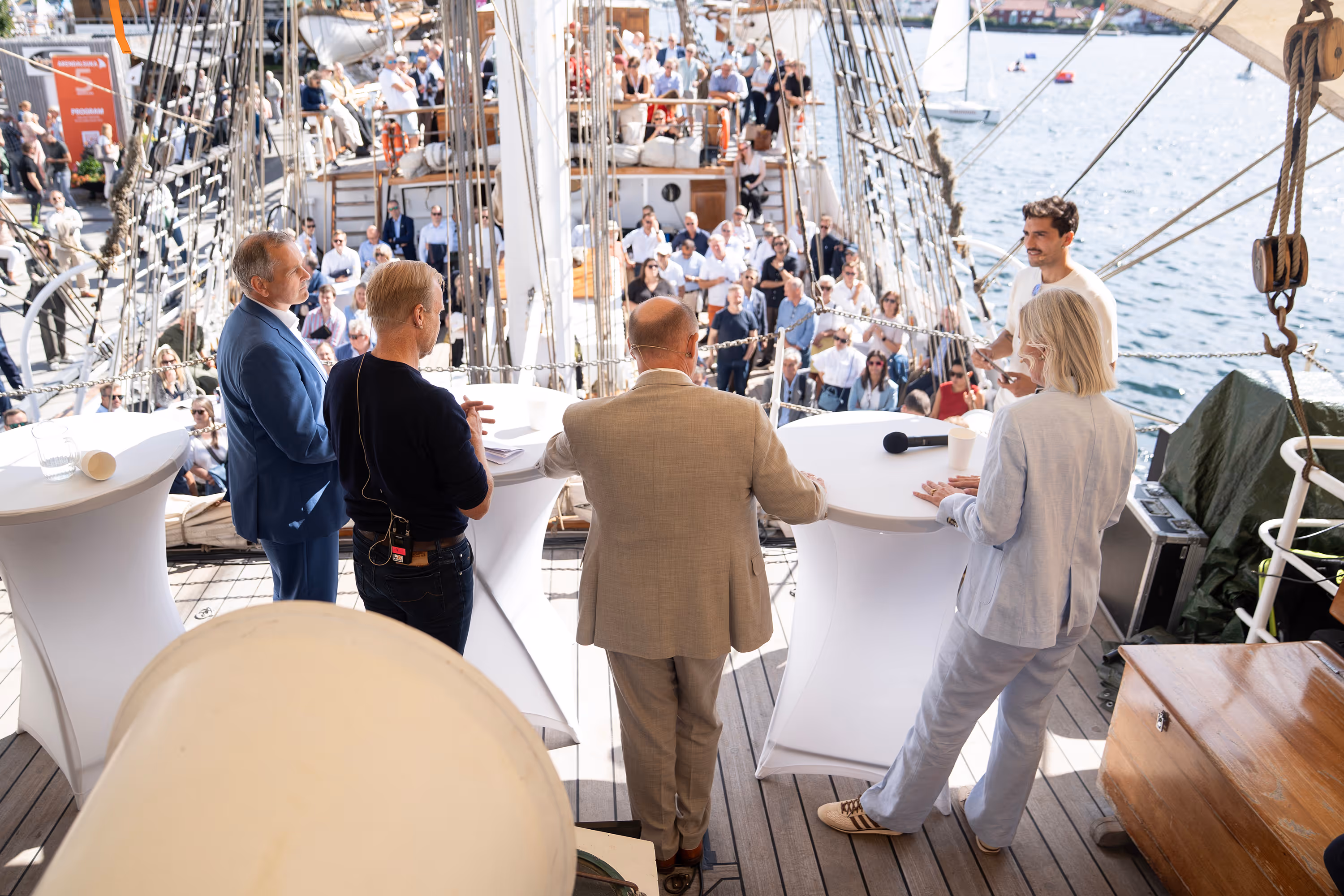 Five people standing behind tables speaking to a crowd on a ship deck near water.