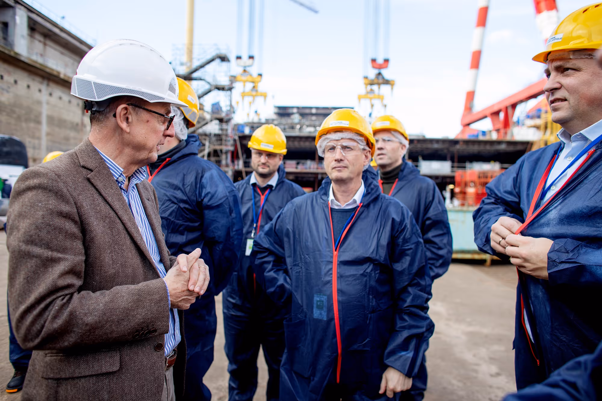 Group of men wearing yellow and white hard hats and blue protective coats at an industrial site.