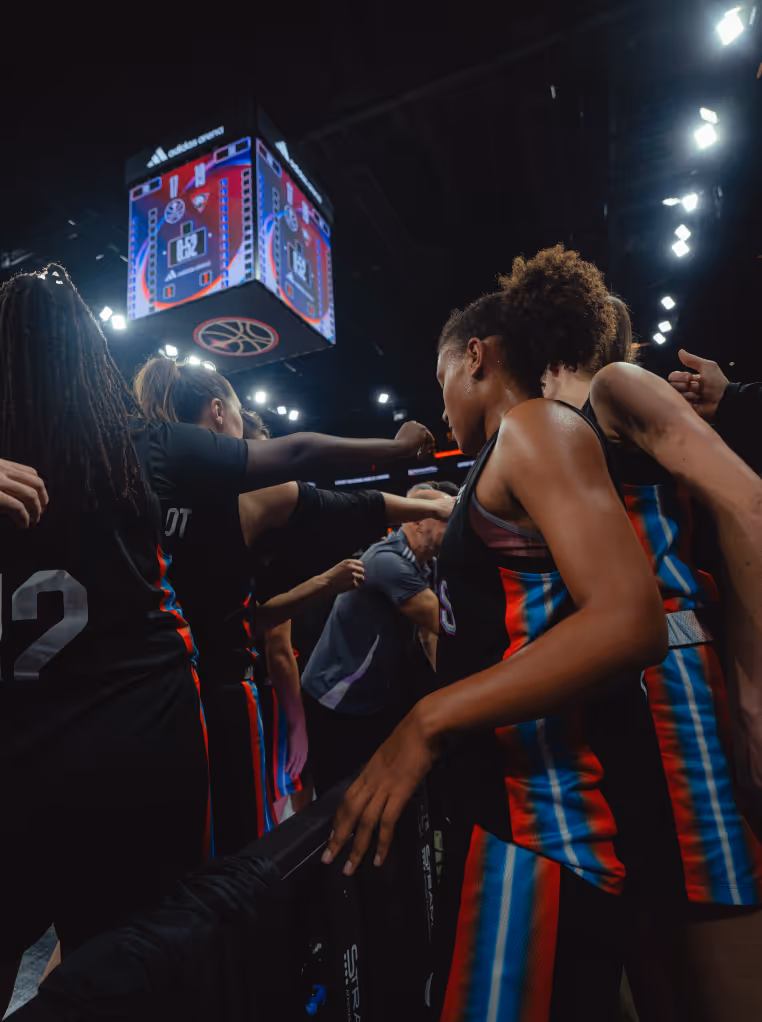 Joueuses de basketball en maillots noirs avec bandes bleues et rouges formant un cercle de motivation sous un tableau de score éclairé.