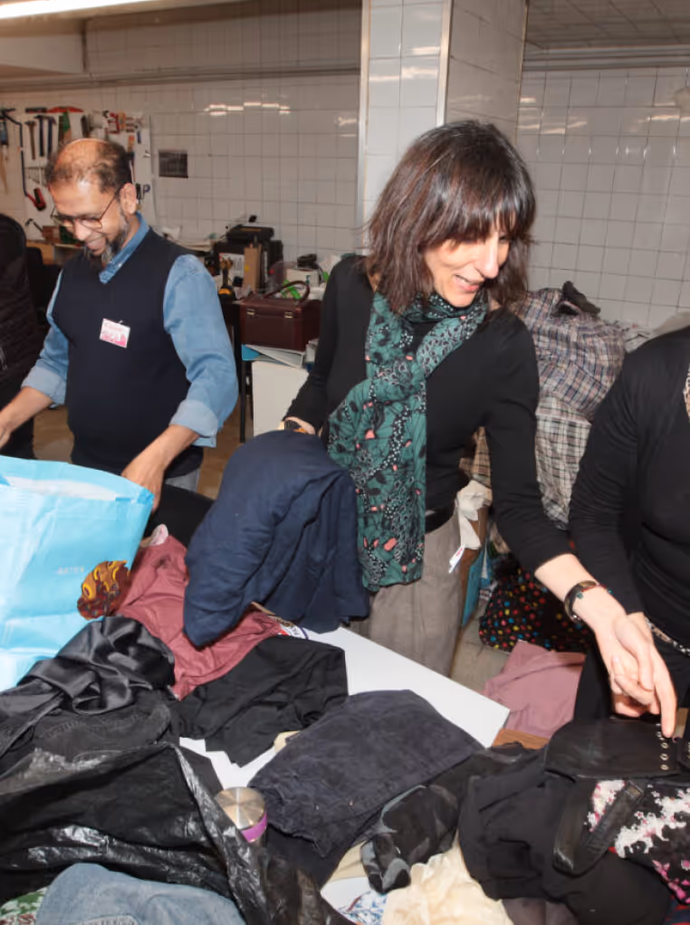 Deux adultes triant des vêtements sur une table dans une pièce intérieure, un homme à gauche souriant et une femme à droite portant une écharpe verte.