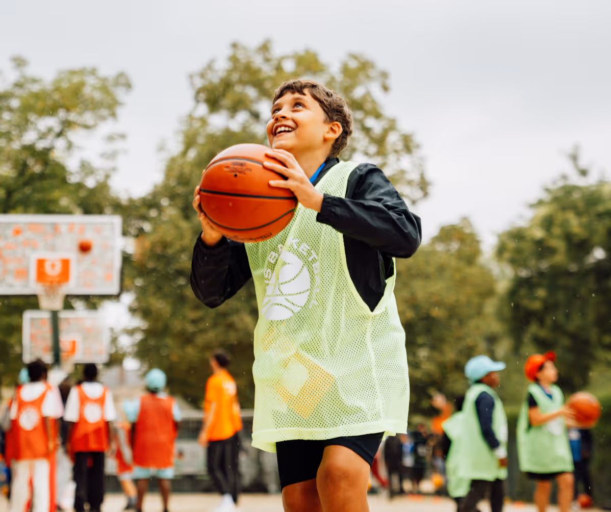 Un garçon souriant en tenue de basket jaune clair tenant un ballon avant de tirer, avec d'autres enfants jouant au basket en arrière-plan sur un terrain extérieur.