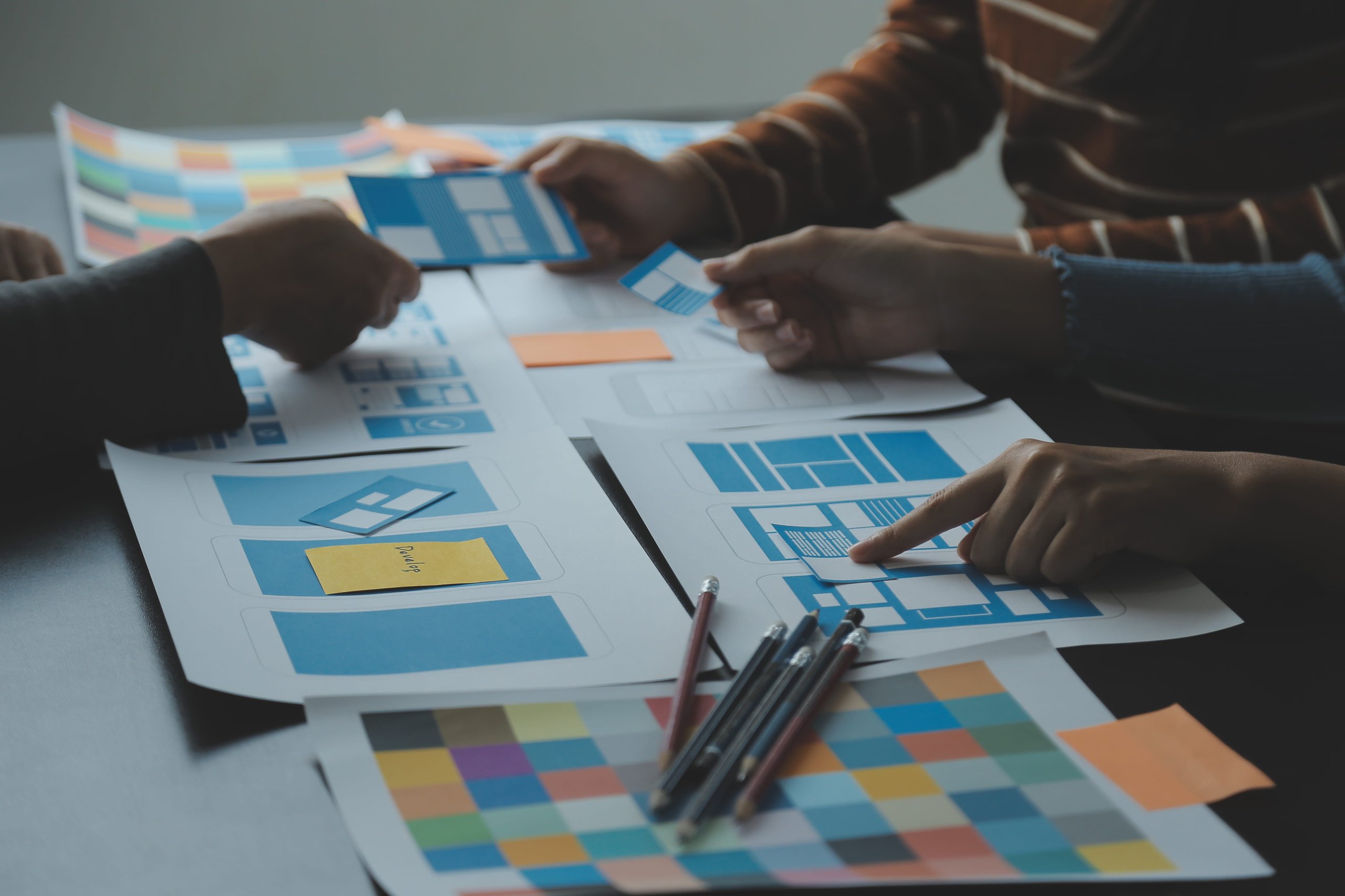 Hands of people collaborating on printed blue and white wireframe designs and color swatches spread on a table.