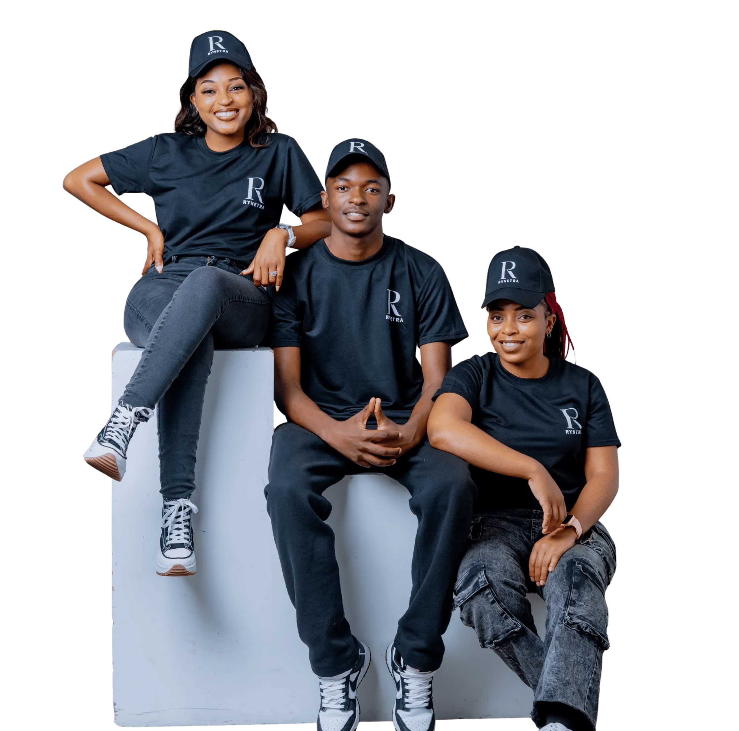 Three young adults wearing black Rynetra caps and shirts, two sitting on a white block and one sitting next to it, smiling.