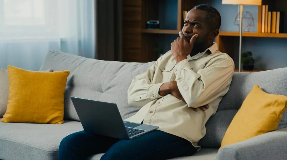 Man sitting on a gray couch with a laptop on his lap, looking thoughtfully to the side with hand on chin.
