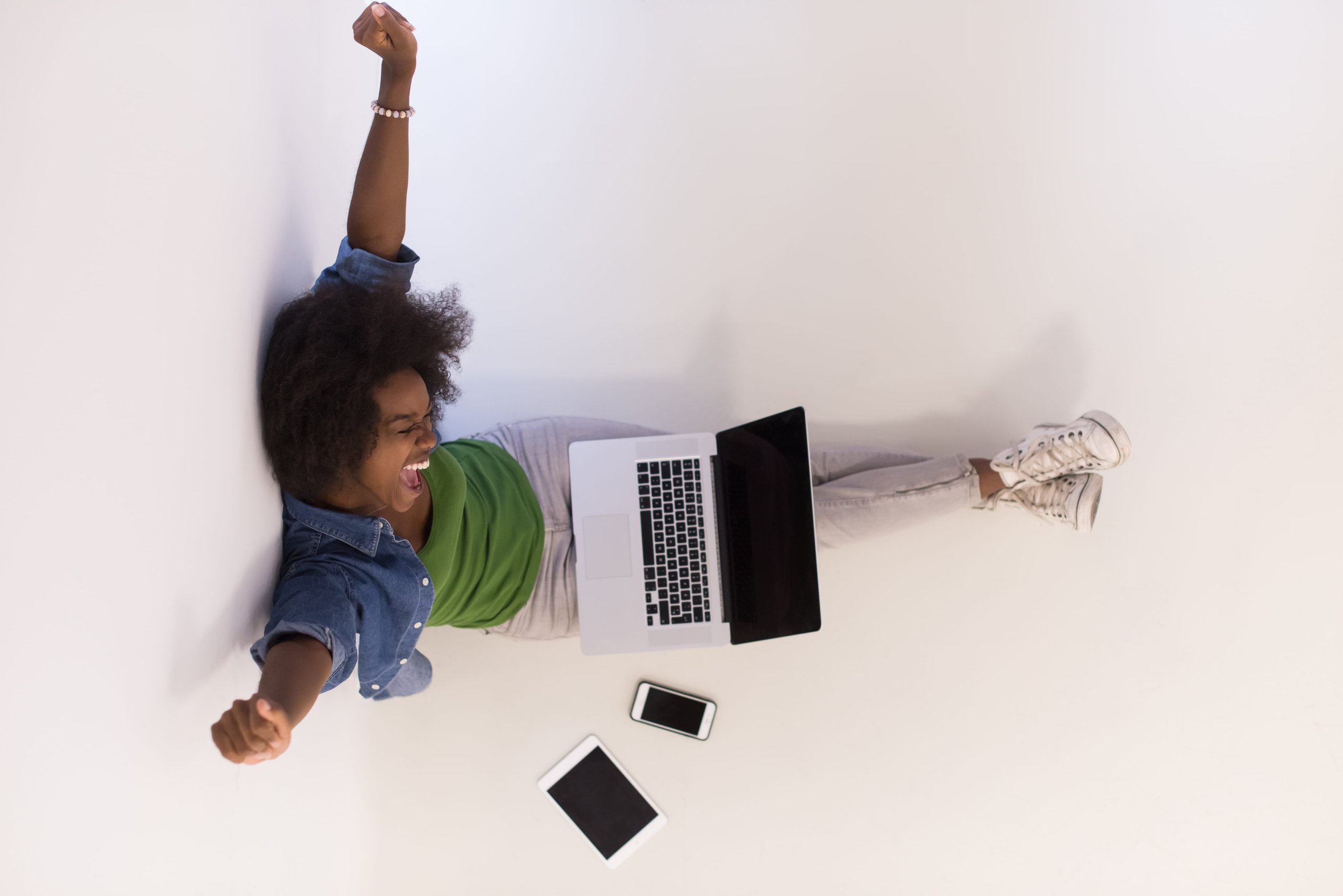 Excited woman stretching arms while sitting on floor with laptop on lap and tablet and smartphone nearby.