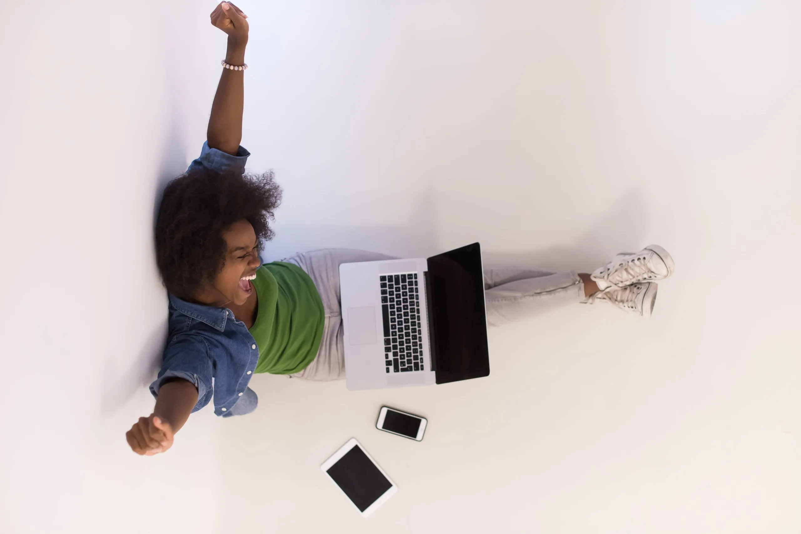 Excited woman stretching arms while sitting on floor with laptop on lap and tablet and smartphone nearby.