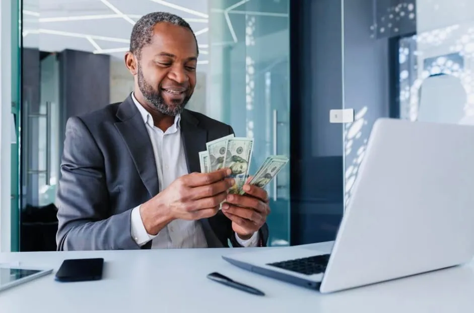 Smiling man in a suit counting US dollar bills at a desk with a laptop, phone, and pen in a modern office.