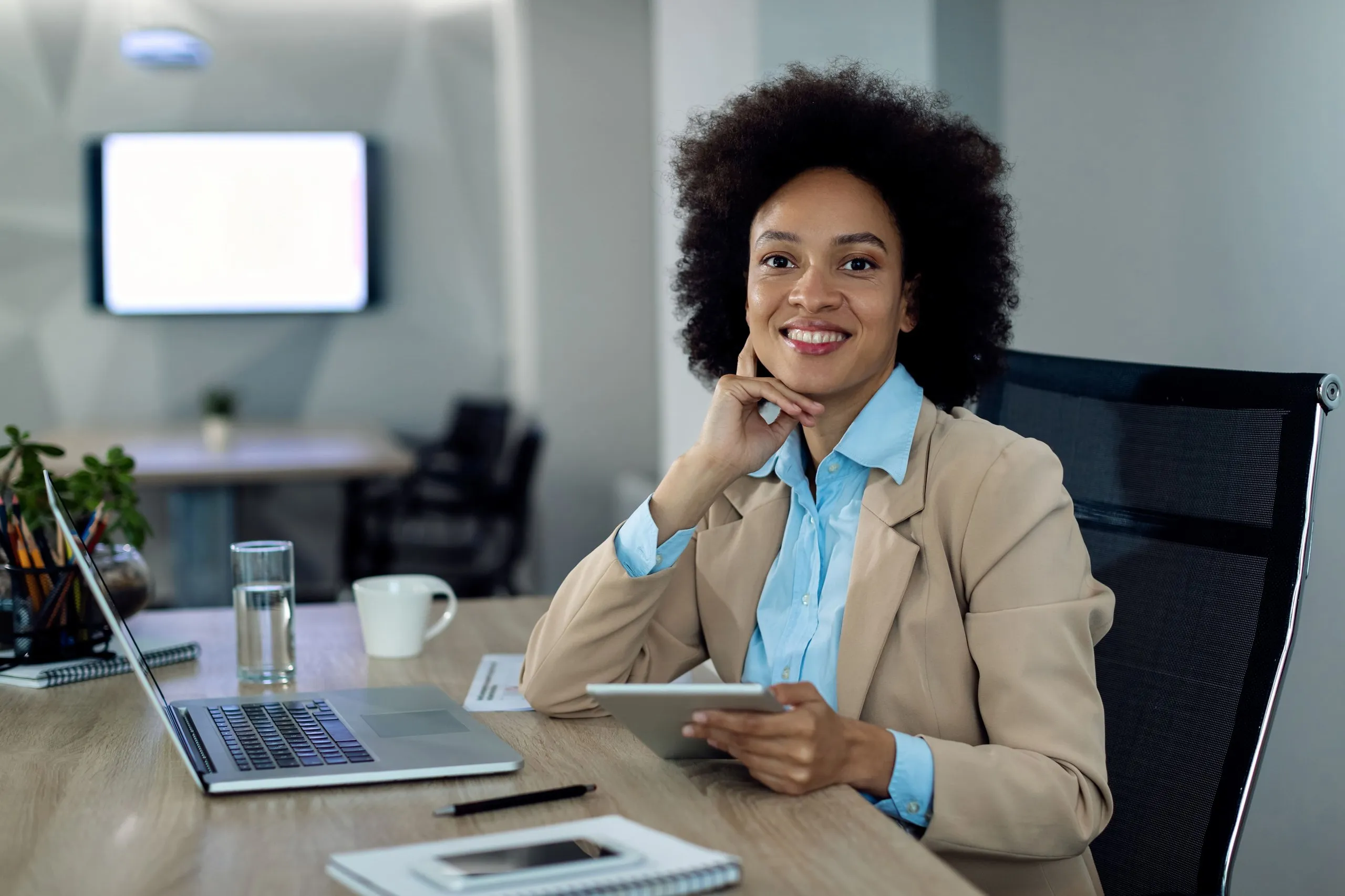 Smiling businesswoman sitting at a desk with a tablet, laptop, and office supplies in a modern workspace.