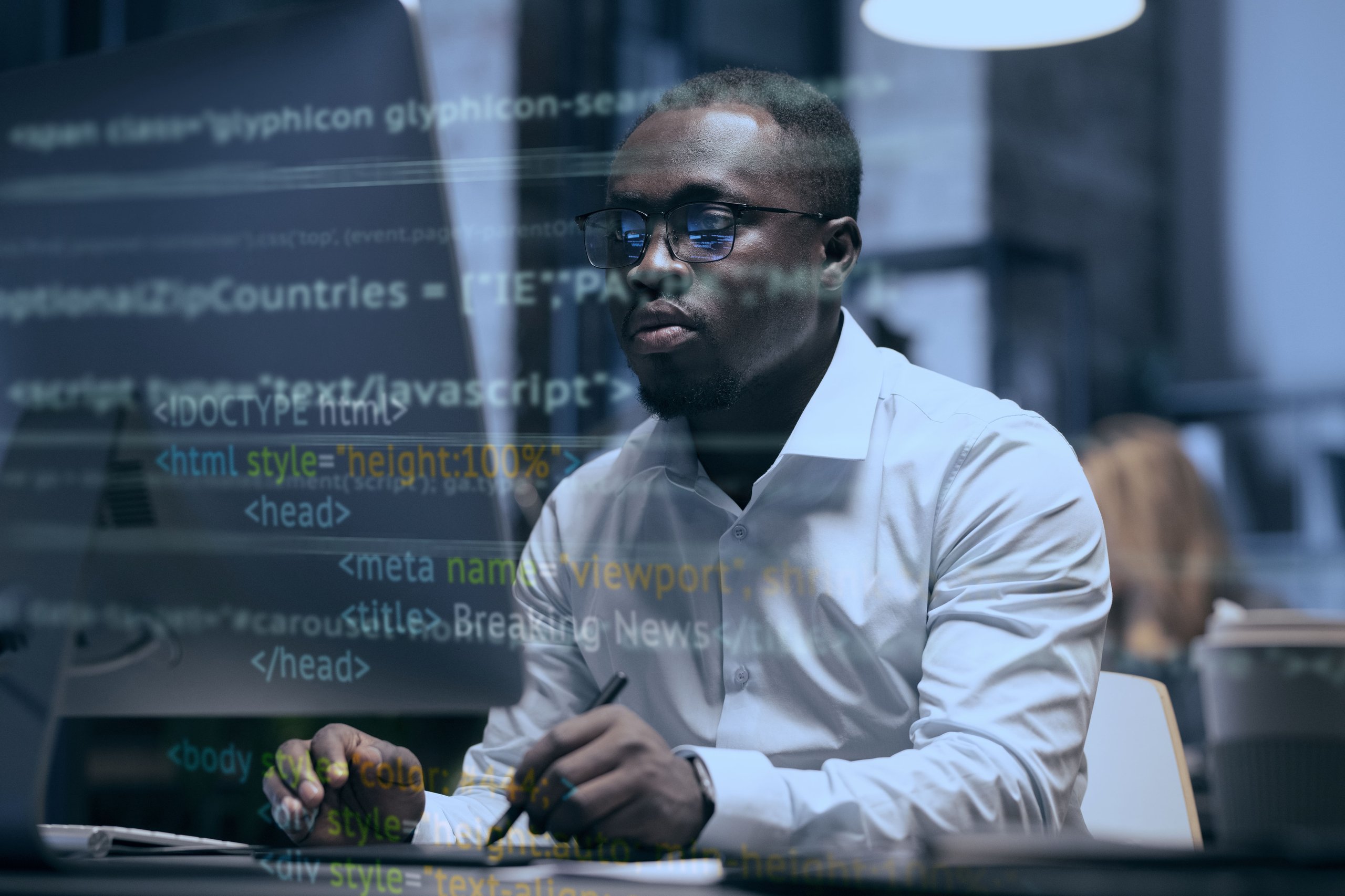 Man in glasses and white shirt working on a computer with HTML code reflected on the screen.