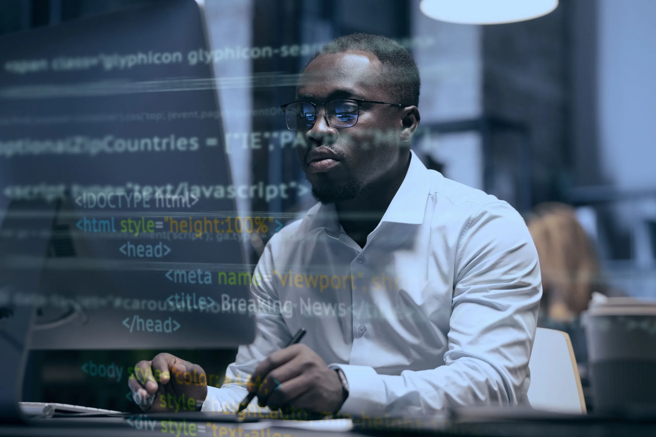 Man in glasses and white shirt working on a computer with HTML code reflected on the screen.