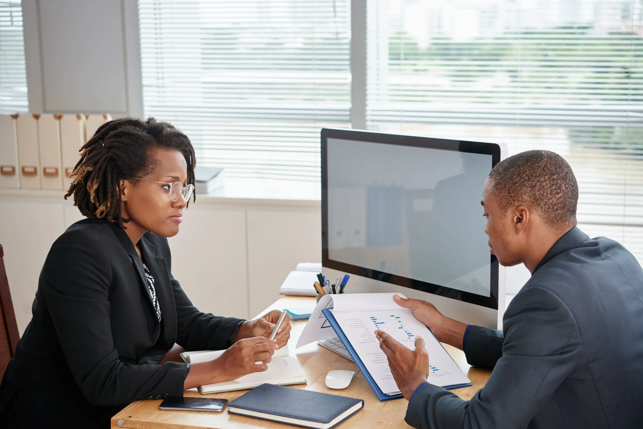 Two professionals reviewing charts together at a desk with a computer in a bright office.