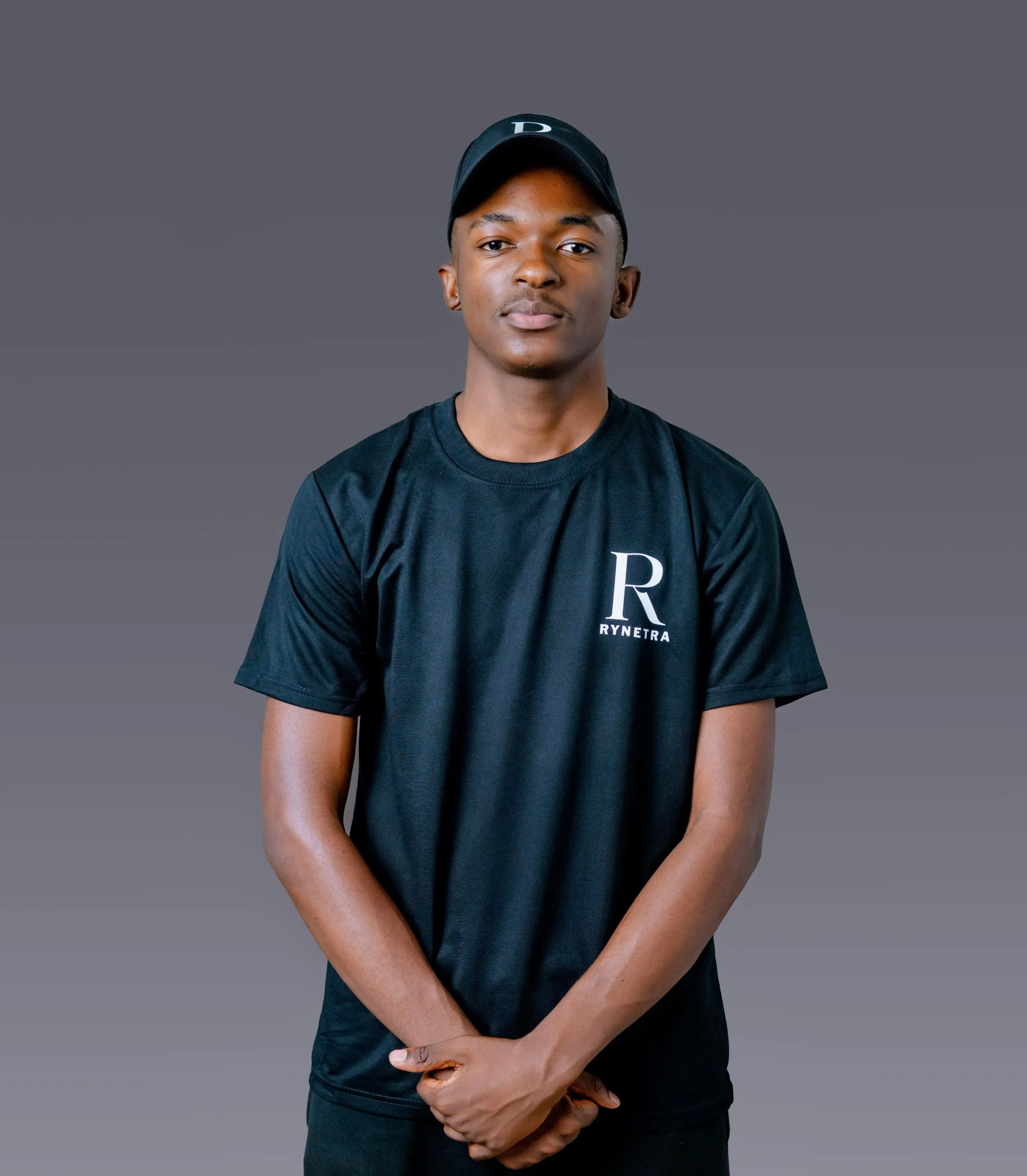 Young man wearing a black Rynetra t-shirt and matching black cap, standing against a plain gray background.