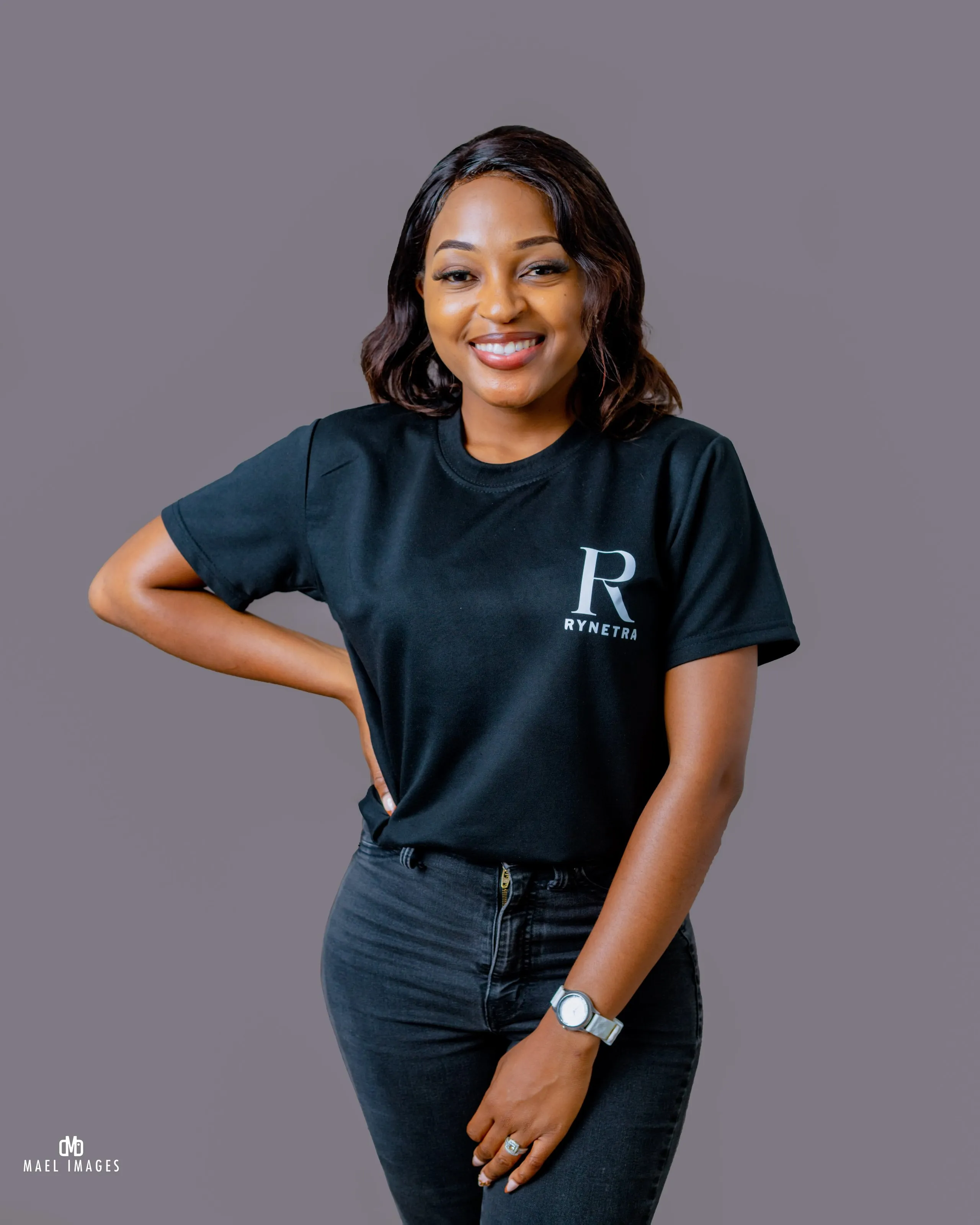 Smiling young woman wearing a black Rynetra t-shirt and dark jeans, posing with one hand on her hip.