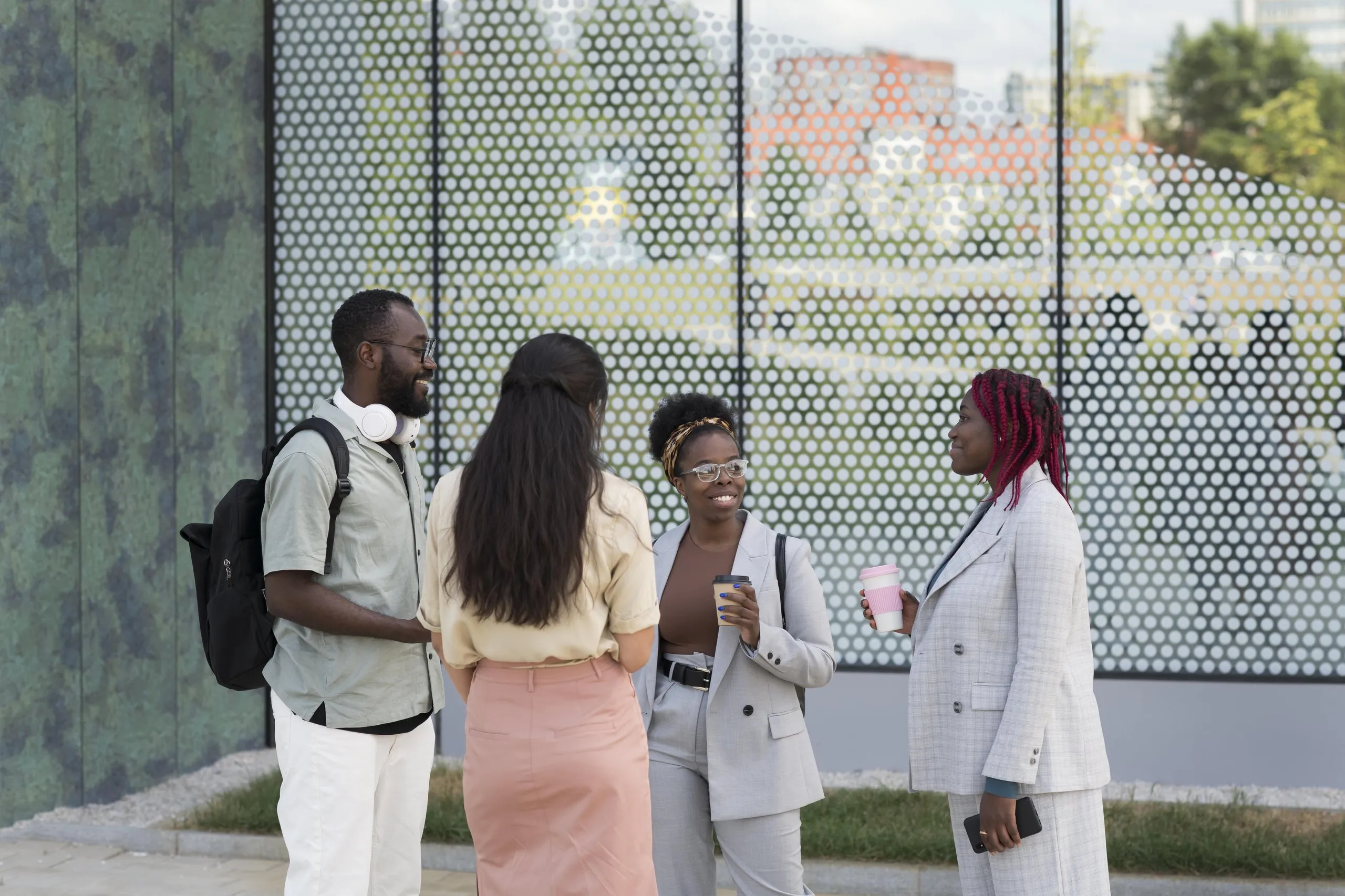 Four young professionals standing outdoors near a modern building, chatting and holding coffee cups.