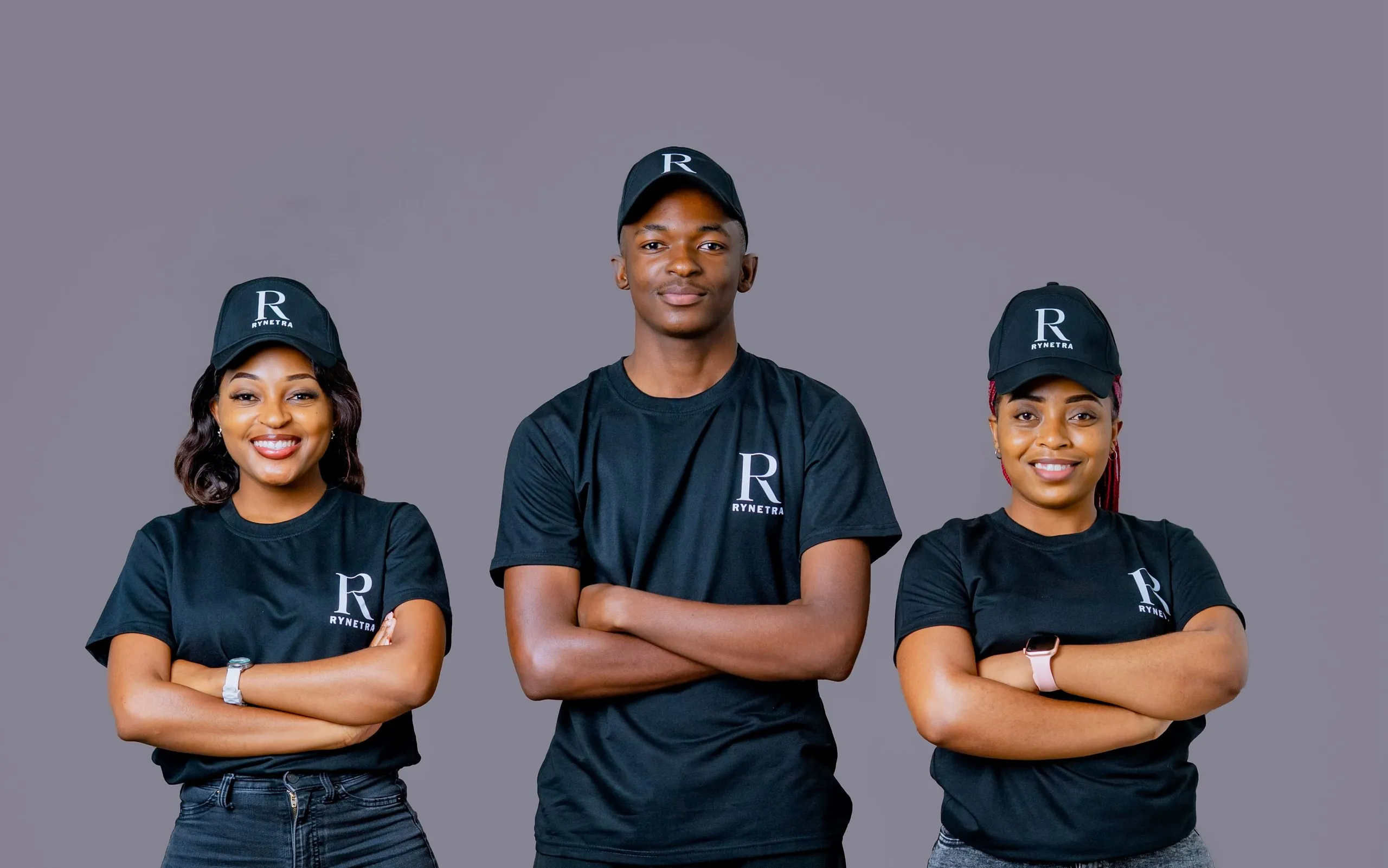 Three people standing side by side against a gray background, wearing black RYNETRA branded t-shirts and caps, smiling with arms crossed.