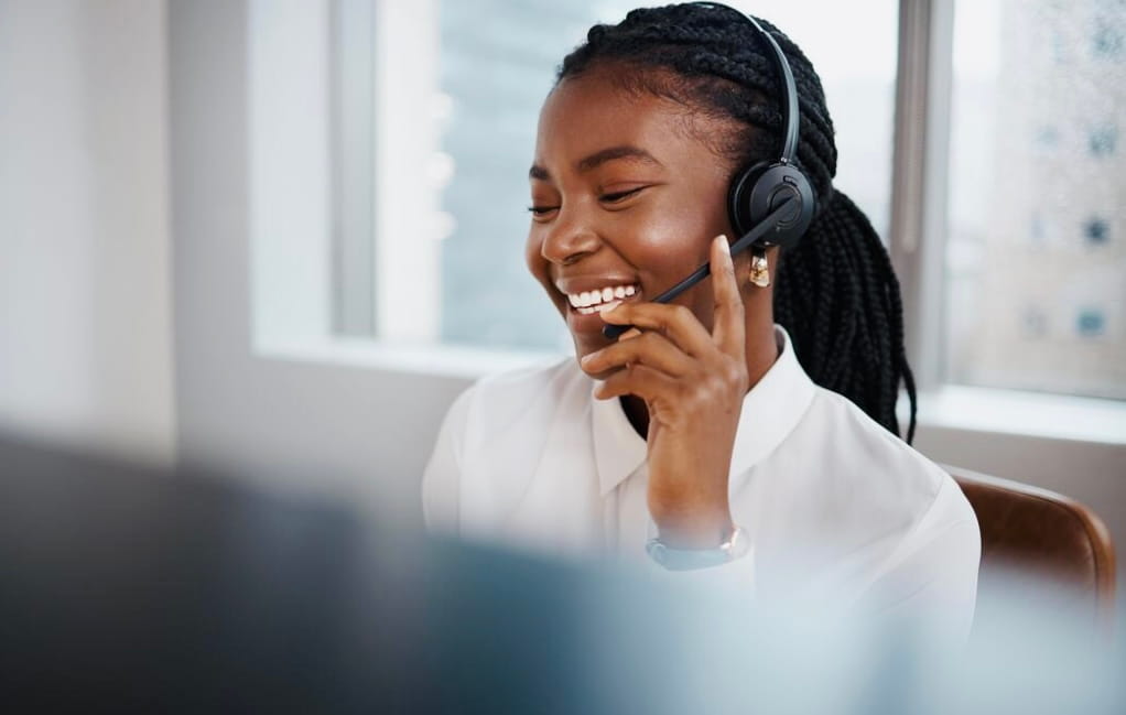 Smiling woman wearing headset with microphone, appearing in a customer service or call center setting.