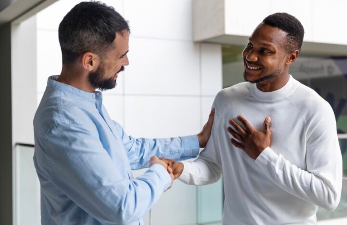 Two men smiling while shaking hands, one with a hand on the other's shoulder in a friendly gesture.