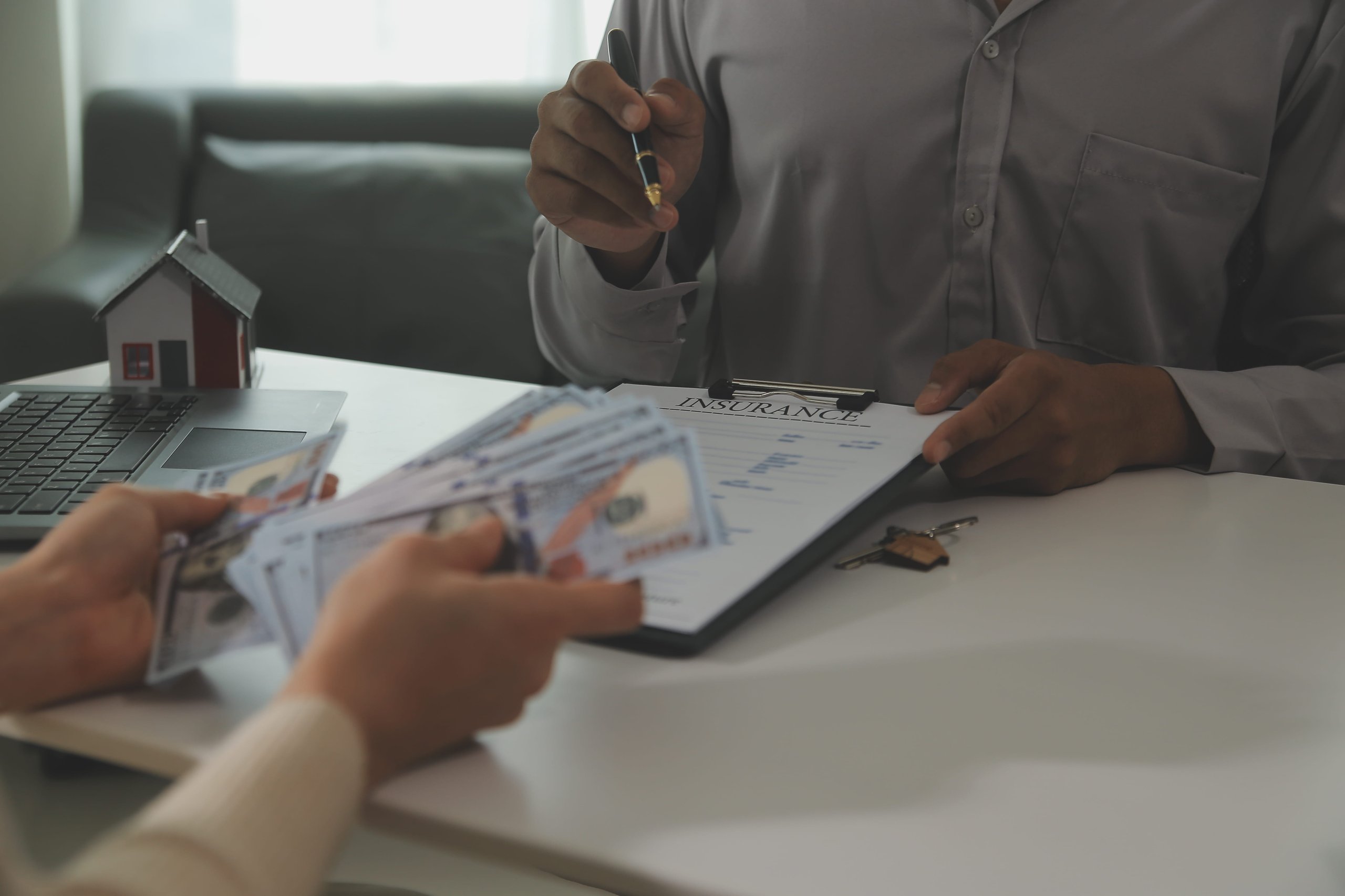 Person holding a pen and an insurance document while another person hands over a stack of US dollar bills across a table with a small house model and a laptop.