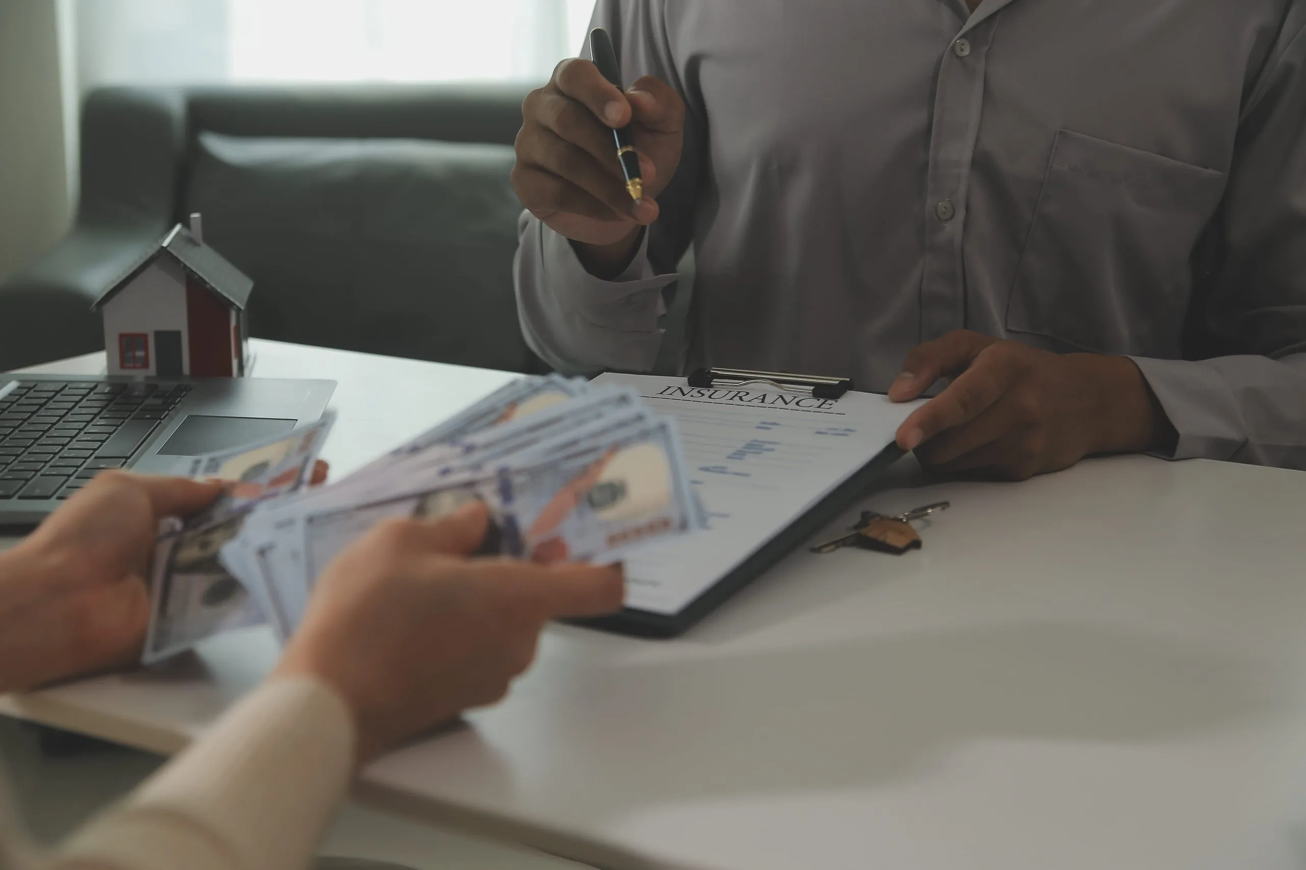 Person holding a pen and an insurance document while another person hands over a stack of US dollar bills across a table with a small house model and a laptop.