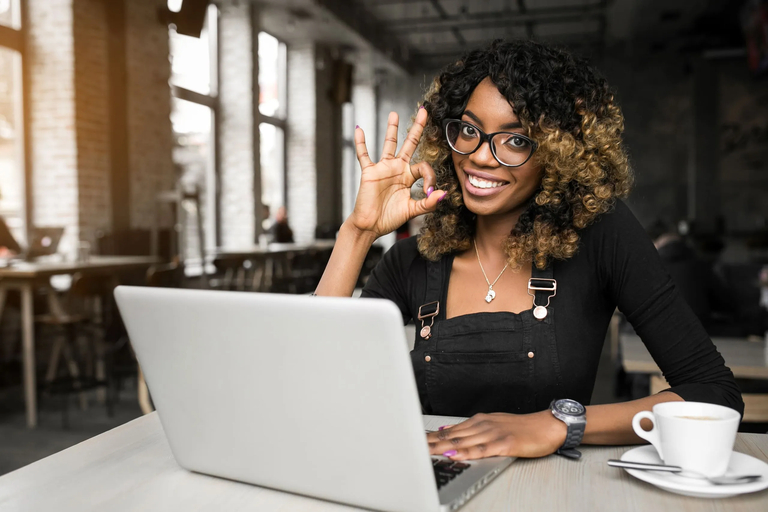 Smiling woman with glasses showing an OK hand sign while working on a laptop in a bright café.