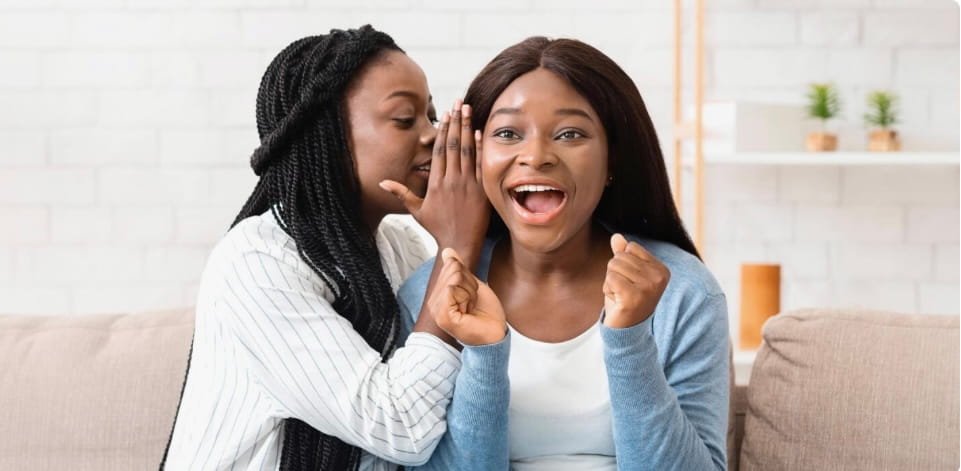 Two women sitting on a couch, one whispering a secret to the other who looks surprised and excited.