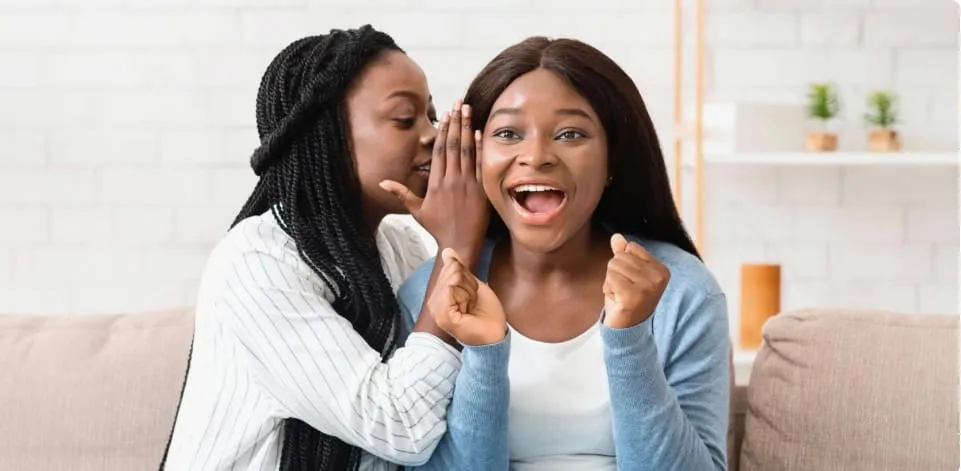Two women sitting on a couch, one whispering a secret to the other who looks surprised and excited.