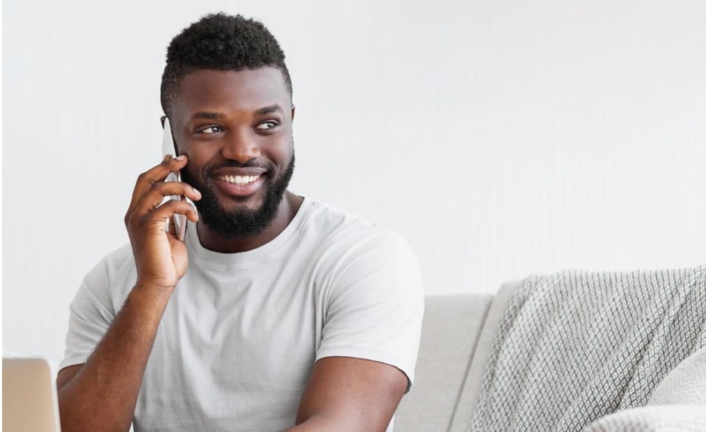 Smiling man in a white t-shirt talking on a smartphone while sitting on a couch with a gray blanket.