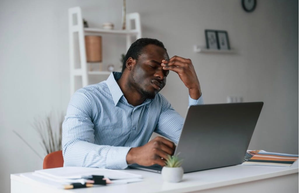 Tired man in a checkered shirt sitting at a desk with a laptop, resting his forehead with his hand.