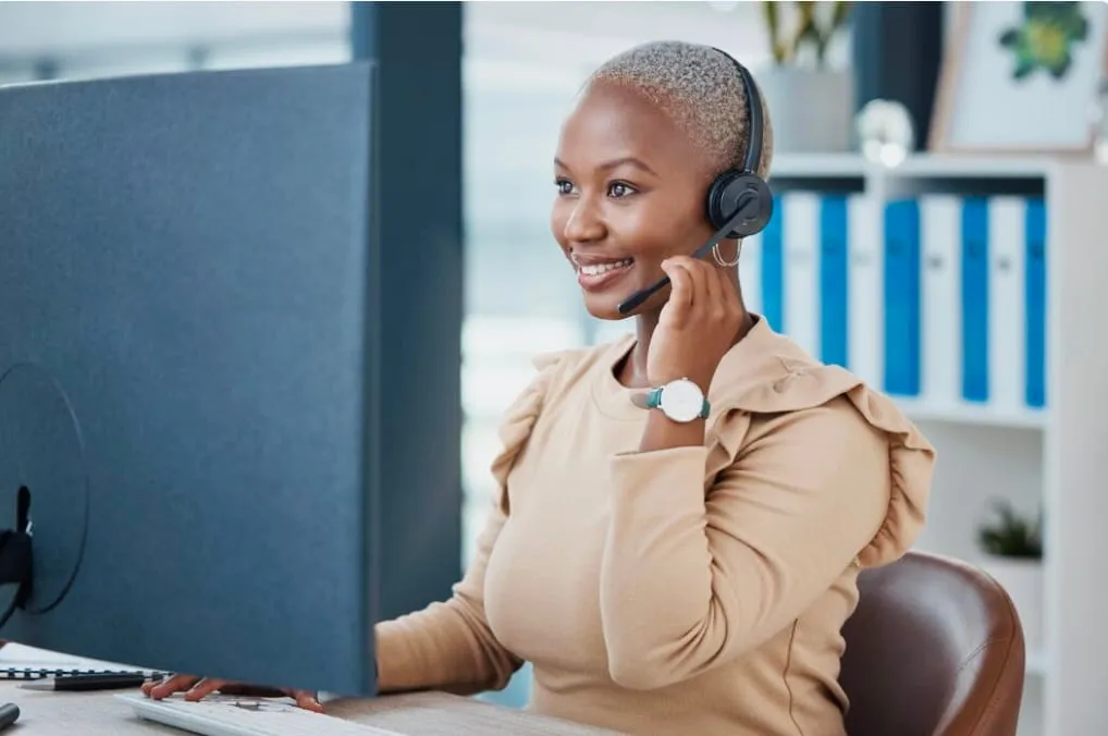 Smiling woman wearing headset and beige top working at a computer in an office.