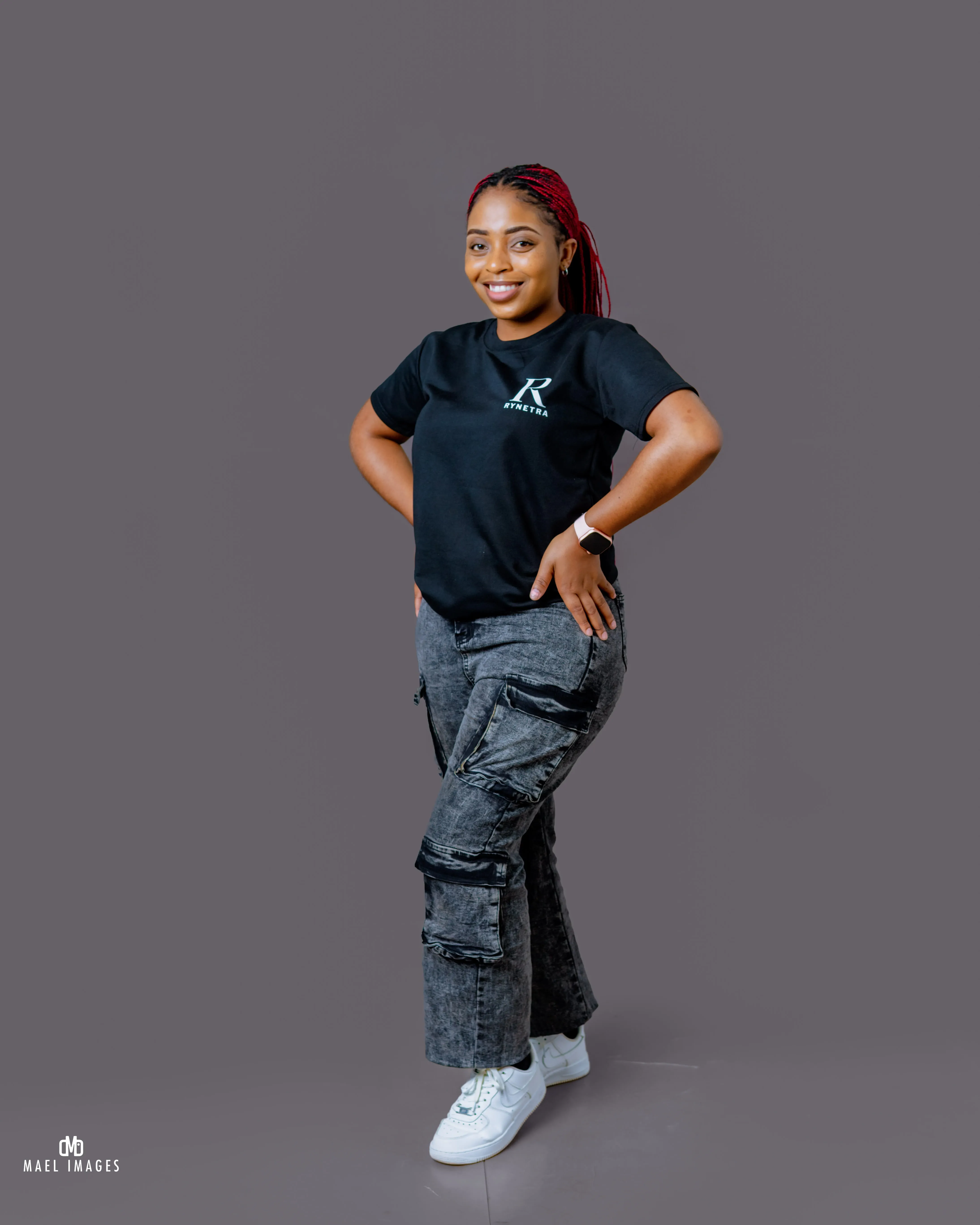 Smiling woman with red braided hair wearing a black Rynetra t-shirt, gray cargo pants, and white sneakers standing with hands on hips.