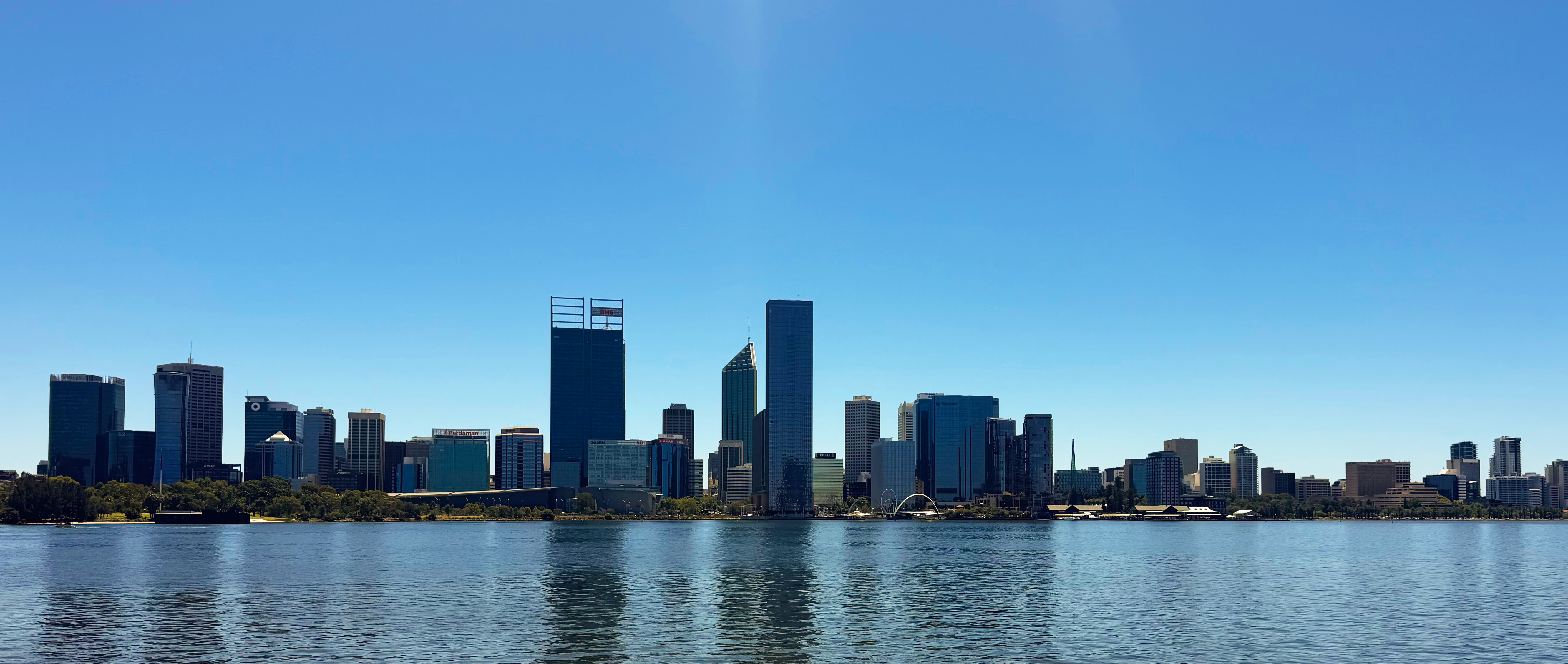Panoramic view of a city skyline with tall buildings reflecting on calm water under a clear blue sky.