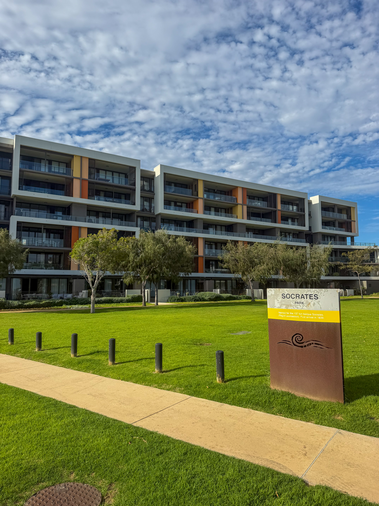 Modern houses with large windows and balconies facing a green lawn and a walkway under a cloudy sky.