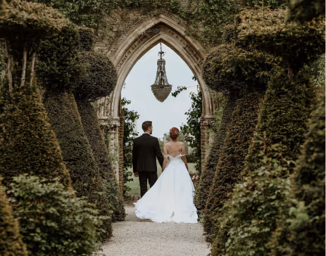 Bride and groom walking hand in hand through a garden archway surrounded by neatly trimmed greenery.