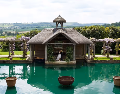 Couple embracing under a thatched gazebo surrounded by a turquoise pool with a green landscape in the background.
