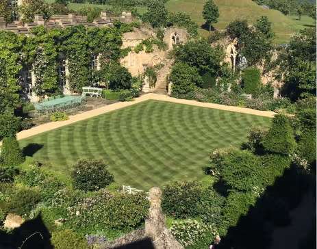 Manicured garden lawn with circular mowing patterns, surrounded by trimmed bushes, ivy-covered stone walls, outdoor table and benches.